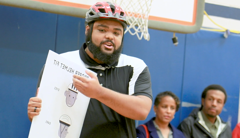 PE teacher speaking and holding sign that says "proper helmet fit."